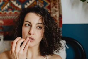 A woman with curly hair applies lip balm in a cozy indoor setting.