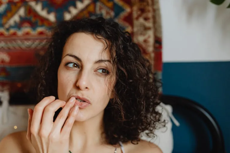 A woman with curly hair applies lip balm in a cozy indoor setting.